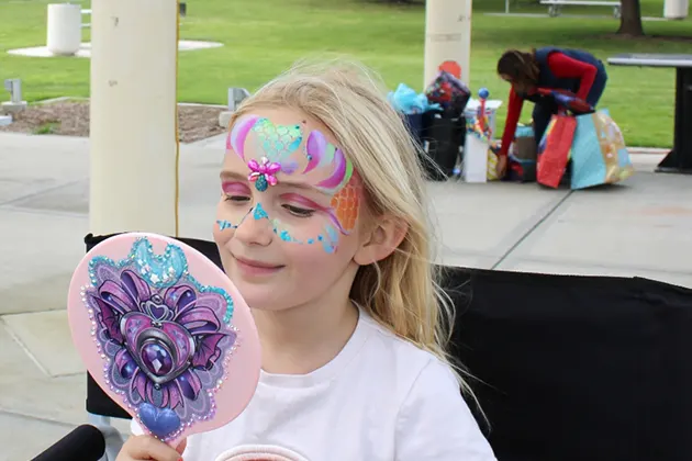 Child with colorful face paint holding a decorated mirror, outdoors.