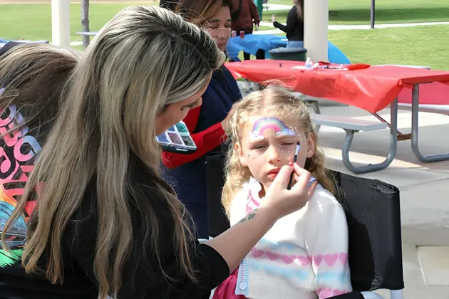 Woman painting a rainbow on a young girl's face outdoors.