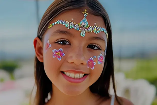 Girl with jeweled face art and floral cheek designs smiling outdoors.