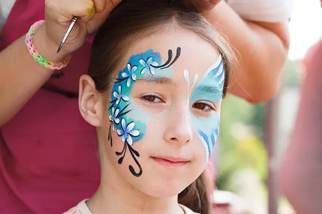 Young girl with blue face paint and flower designs being painted.
