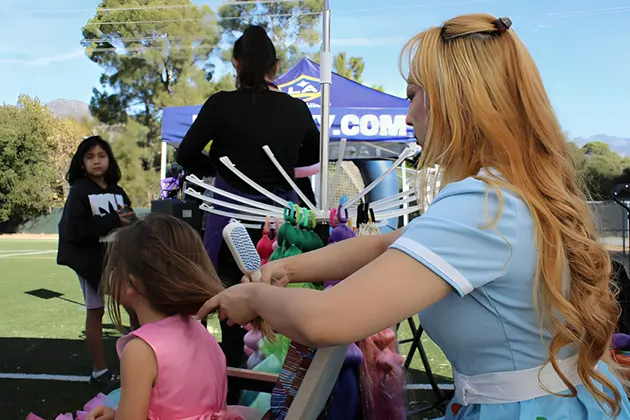 Woman styling girl's hair outdoors with others nearby.